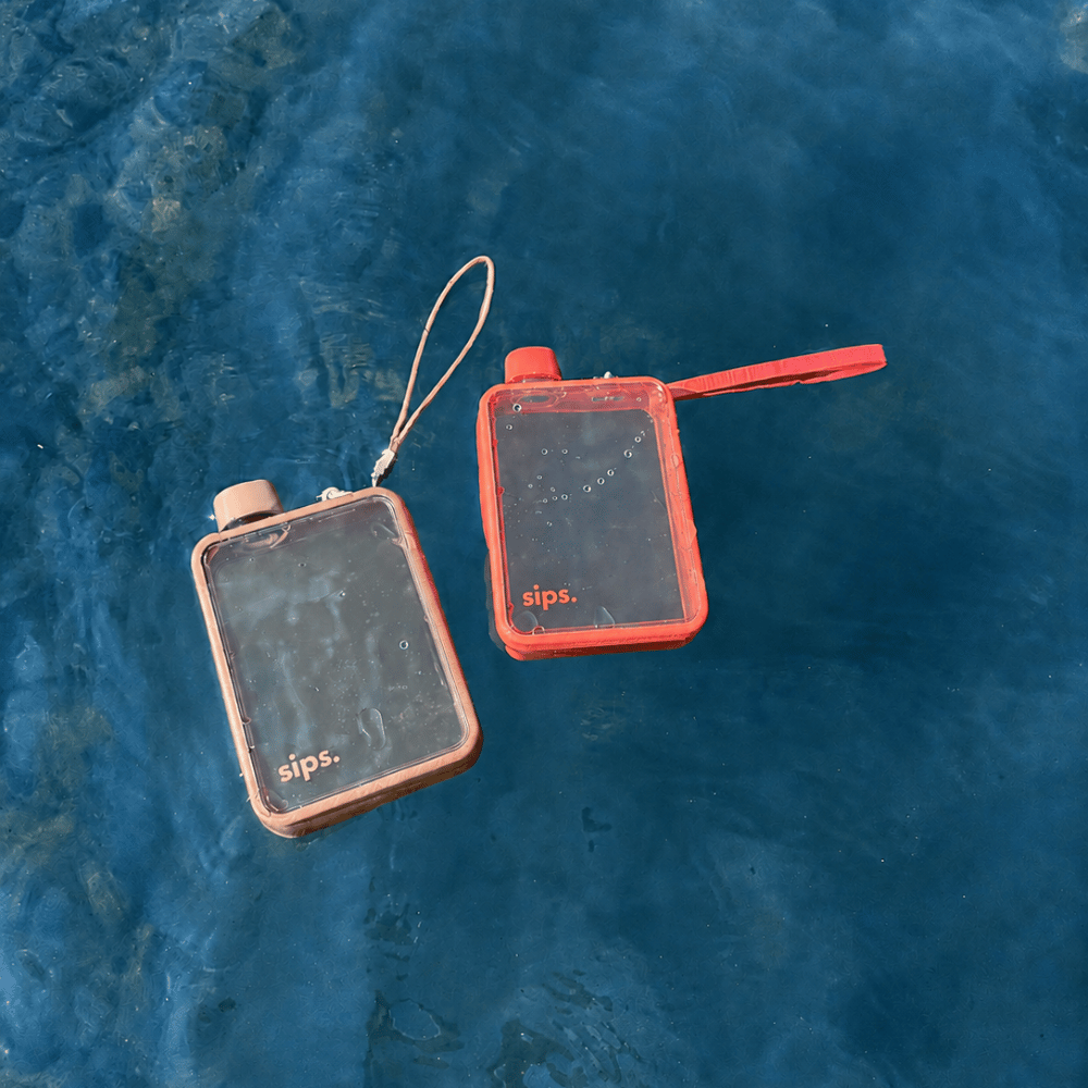 Two clear flask shaped water bottles made by sips in Coral and Taupe floating in blue waters. The perfect water bottle to take on holiday.