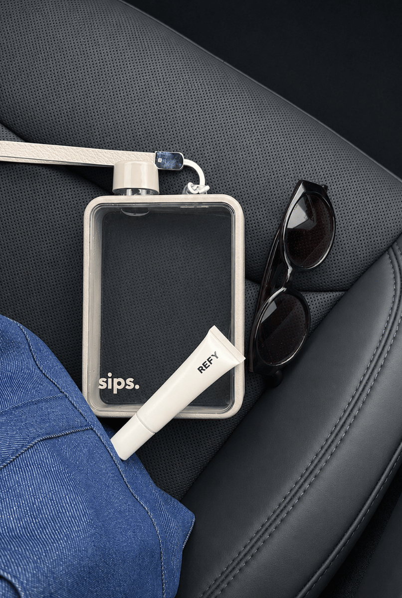 A clear rectangular water flask made by Sips with a light grey bumper and cap, resting on a black car seat alongside black sunglasses, a blue denim jacket, and a white tube labeled REFY. 