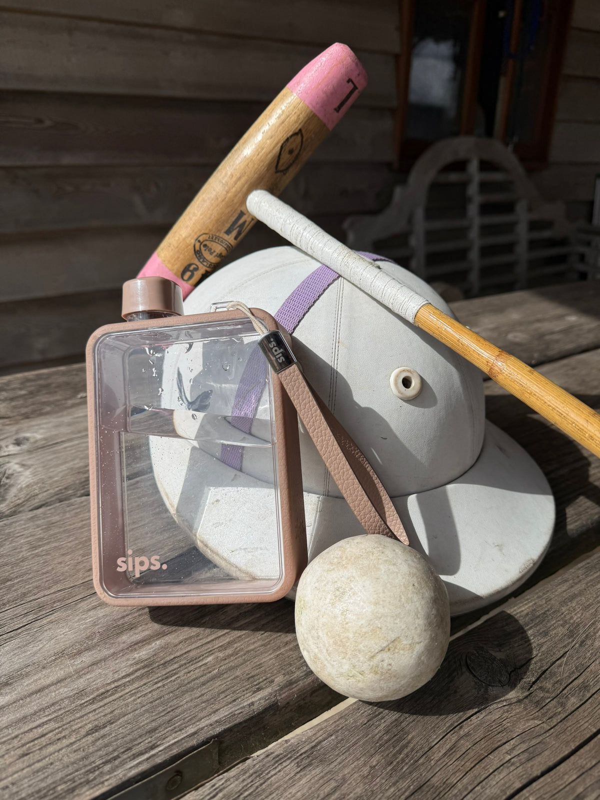 Sips water bottle in taupe set on a wooden table with a polo stick, polo hat and polo ball. The perfect water bottle for polo players.
